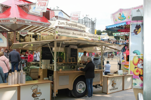 Ein Verkaufsstand mit bunten Schirmen, der Snacks und Getränke anbietet. Viele Menschen im Hintergrund.